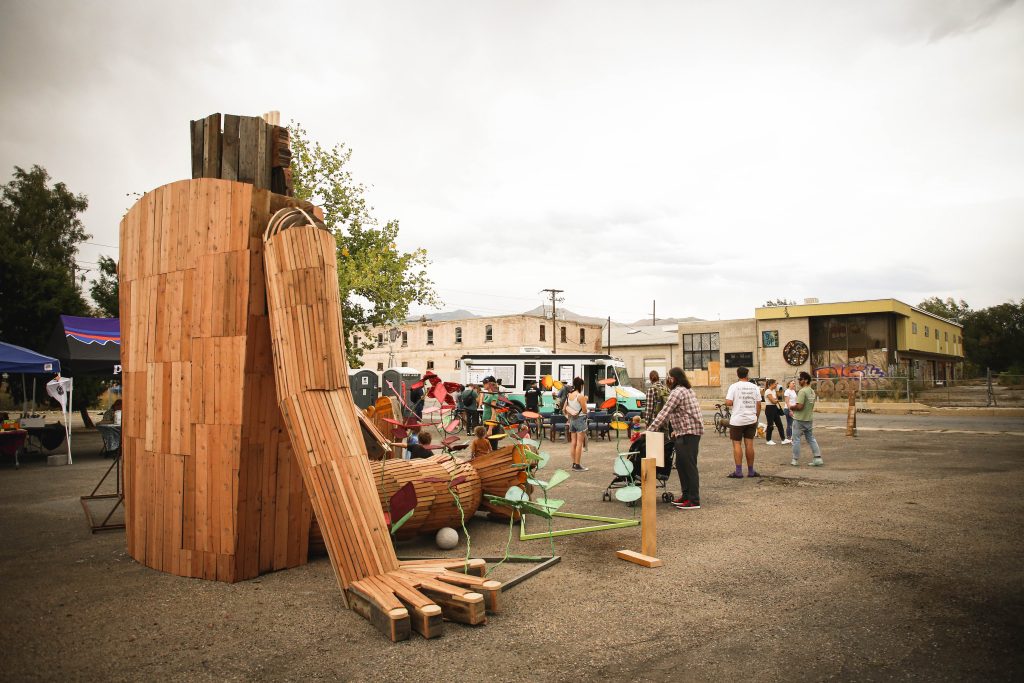 Outdoor scene in the Granary District with a large wooden sculpture and people gathered around in a paved area, with buildings and tents in the background.