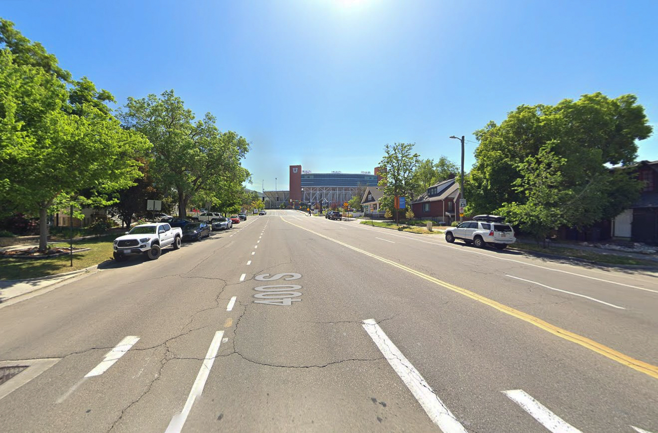 The image shows a street view of a road labeled "400 S" leading toward the University of Utah stadium in Salt Lake City. The street has two lanes with a dividing line, and there are vehicles parked along the curb on both sides. Trees line the sidewalk, providing shade, and residential homes are visible on the right side. The large structure of the stadium dominates the background, with its recognizable red "U" logo on a prominent tower. The weather is clear with bright blue skies.