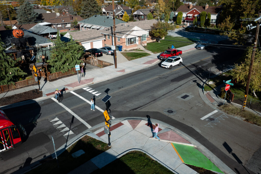 An aerial view of the intersection at Kensington Avenue and 900 East showing pedestrian and bicycle safety improvements. Curb extensions with red paving shorten the crossing distance and slow turning vehicles, while integrated green ramps allow cyclists to easily transition to the sidewalk level. The crosswalk is equipped with two flashing safety signs activated by a push button. On the west side of the street, the sidewalk has been rebuilt extra wide to accommodate cyclists.