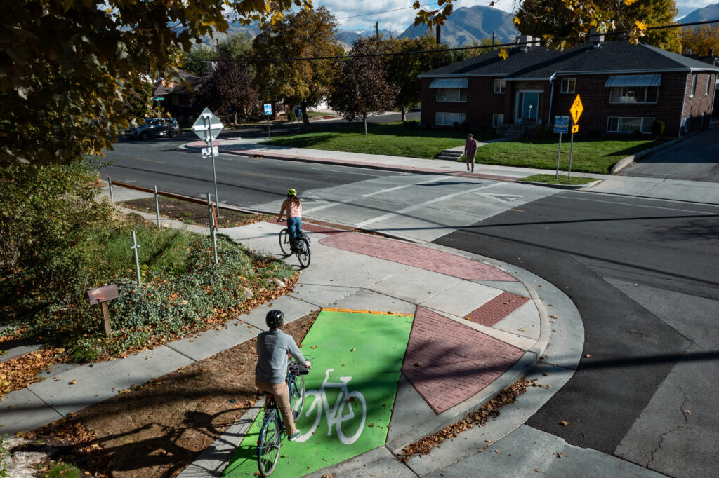 The intersection of the Kensington Avenue Neighborhood Byway and 500 East, where the route jogs north-south. To accommodate the jog, the intersection features a raised crossing that calms traffic and improves ADA access. Curb extensions with red paving narrow the street to slow turning vehicles, while green-painted bike ramps allow cyclists to easily transition from the road to the sidewalk level to utilize the crossing.