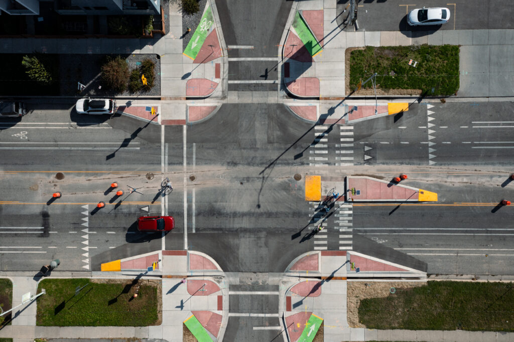 Aerial top-down view of the intersection at Kensington Ave and Main Street, showcasing Kensington neighborhood byway infrastructure designed for safety. The intersection features prominent red-paved curb extensions (bulb-outs) that shorten pedestrian crossing distances, featuring integrated ramps for cyclists. A central pedestrian refuge island with yellow tactile paving sits in the middle of the main roadway.