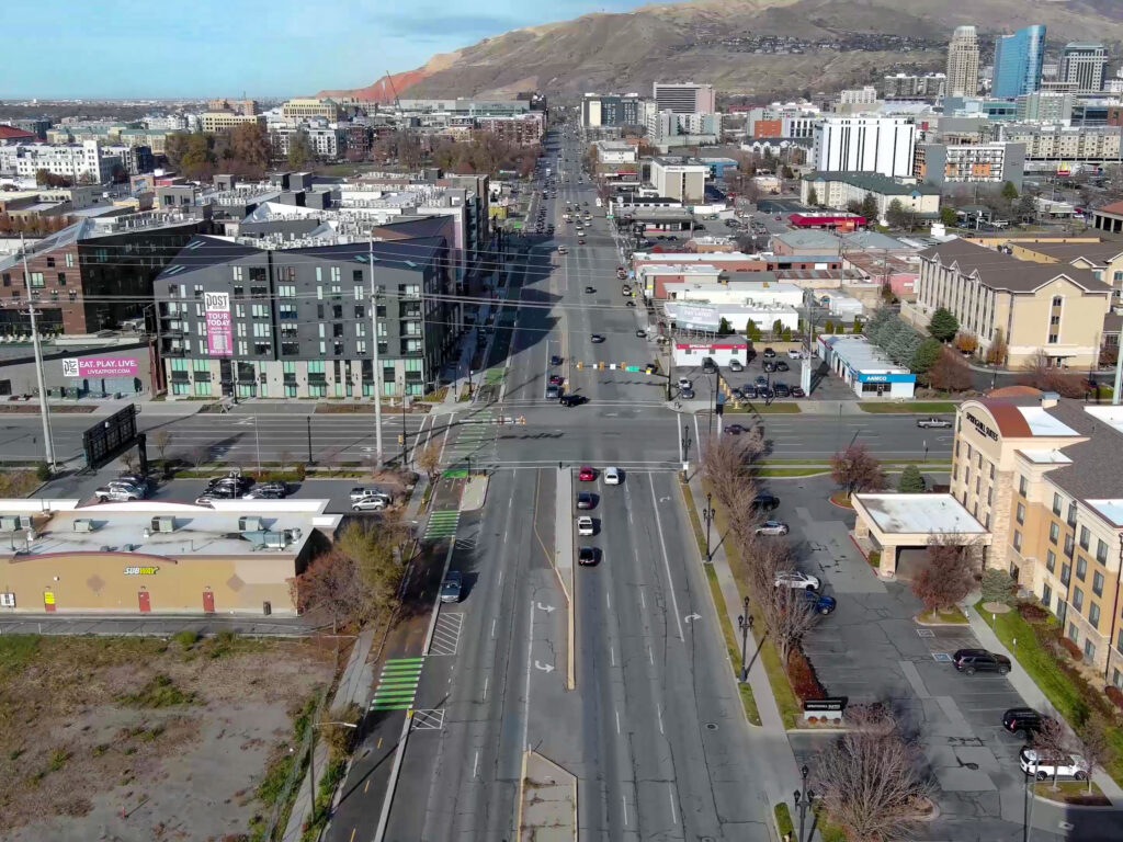 An aerial view of 300 West at 600 South, looking north toward the mountains. A protected two-way bikeway runs along the left side of the street, separated from vehicle traffic by a wide buffer zone. Bright green paint clearly marks the bike path through the intersection and across driveways, creating a continuous, high-visibility route for cyclists.