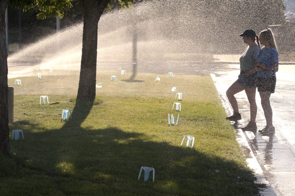 Two women standing on a sidewalk next to a sprinkler watering a lawn.